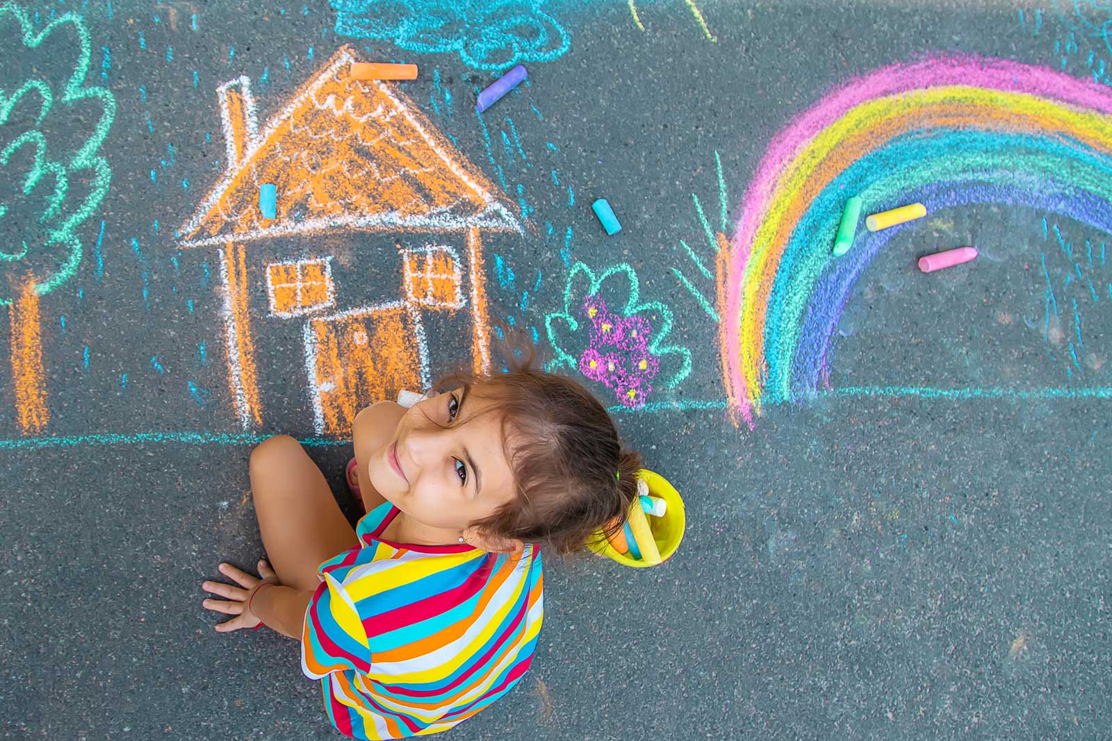 Ein junges Mädchen zeichnet mit bunter Kreide ein Haus und einen Regebogen auf Asphalt.